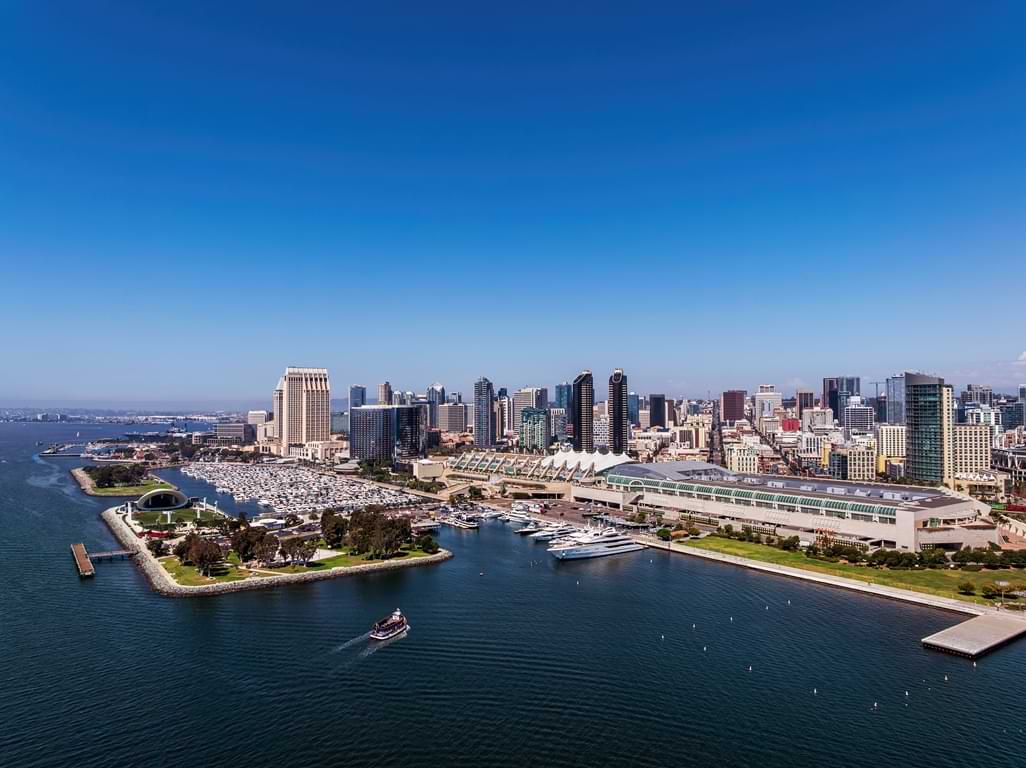 San Diego Convention Center building along the San Diego Bay with the downtown skyline behind and blue skies above.